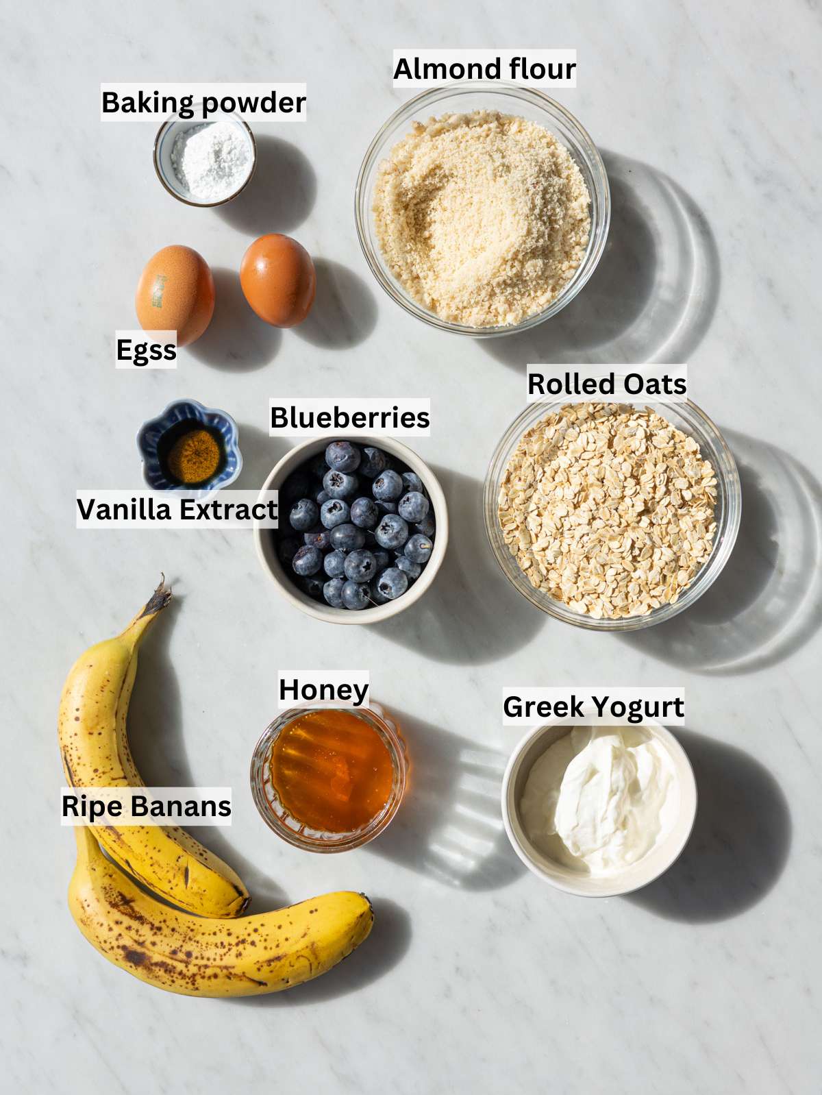 Fresh blueberries, almond flour, rolled oats, and honey arranged on a bright marble surface.