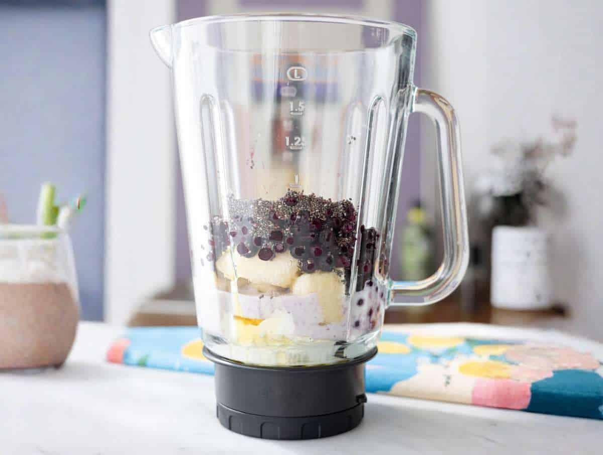 Greek yogurt and milk poured into a blender base next to frozen fruit on marble.
