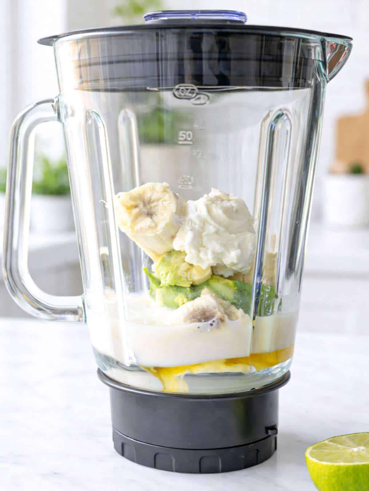 Greek yogurt avocado smoothie ingredients in blender with banana, avocado, yogurt, and milk on marble surface.