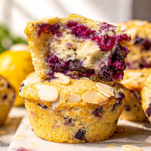 Lemon blueberry muffins stacked on marble table with fresh blueberries and lemon nearby.