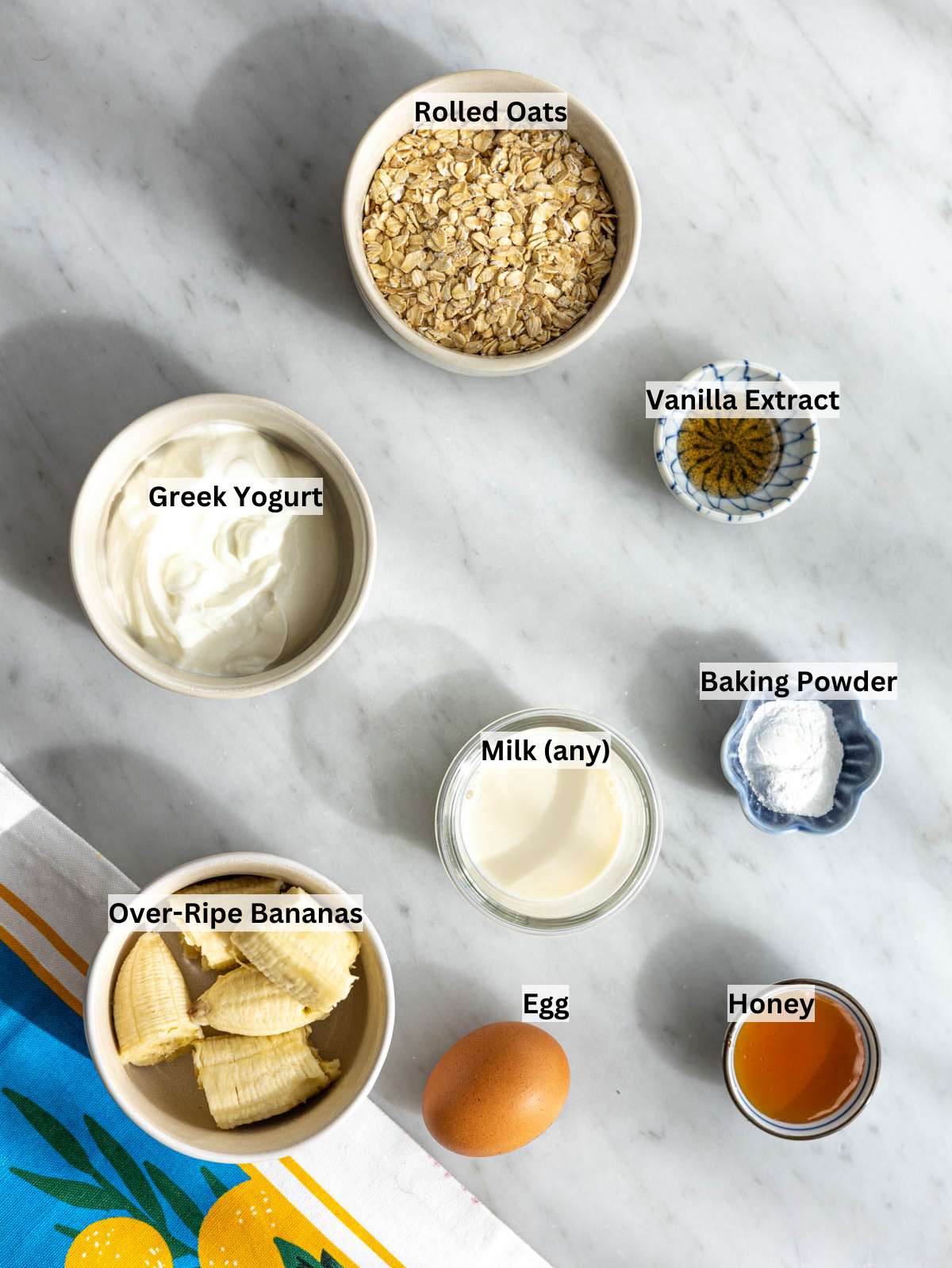 Rolled oats, banana, Greek yogurt, milk, egg, and baking powder on a marble countertop in natural light.