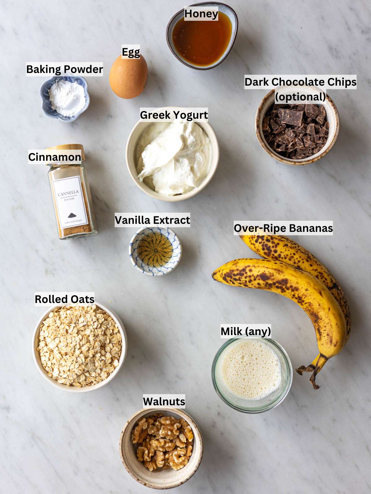 Rolled oats, bananas, Greek yogurt, honey, egg, and walnuts arranged on marble surface in natural light.