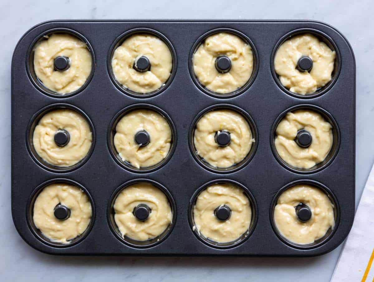 Step 3 Spoon filling donut pan with smooth batter on marble countertop.