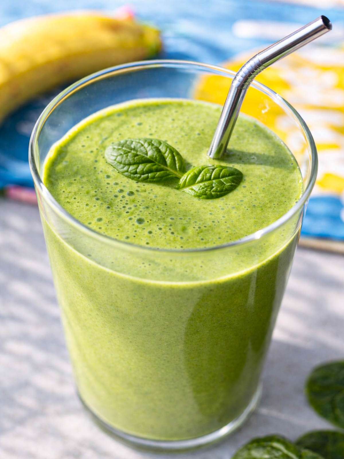 Tall glass of bright green smoothie with spinach leaf and metal straw on marble table.