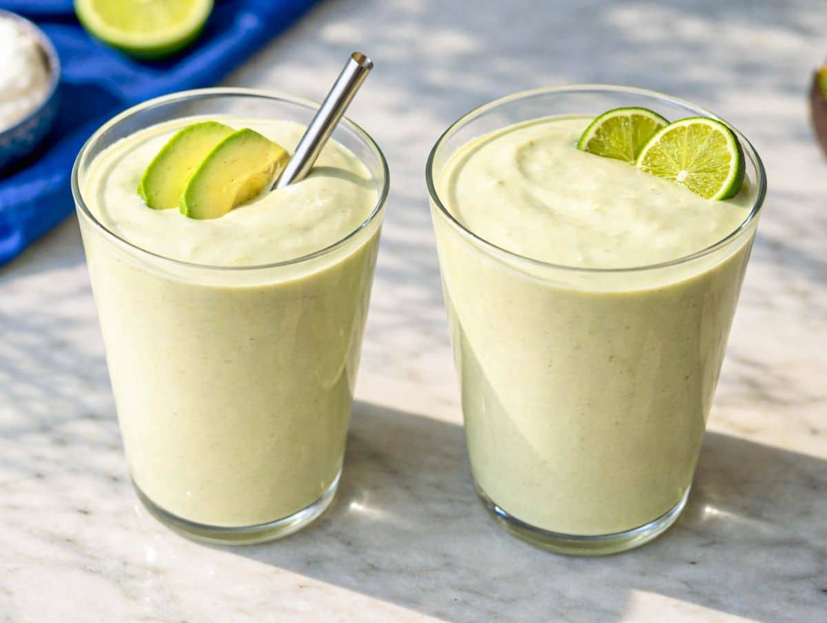 Two glasses of creamy avocado smoothie topped with lime slices and avocado on marble surface.