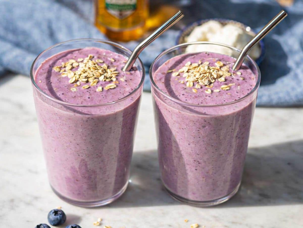 Two glasses of creamy purple Greek yogurt breakfast smoothie topped with oats on marble table in natural light.