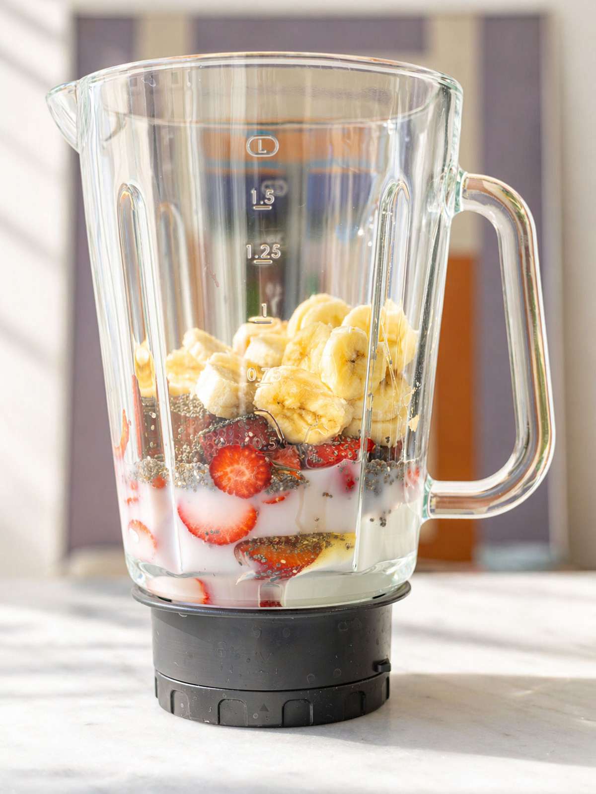 Banana, strawberries, chia seeds, yogurt, and milk layered in blender jar on marble surface.
