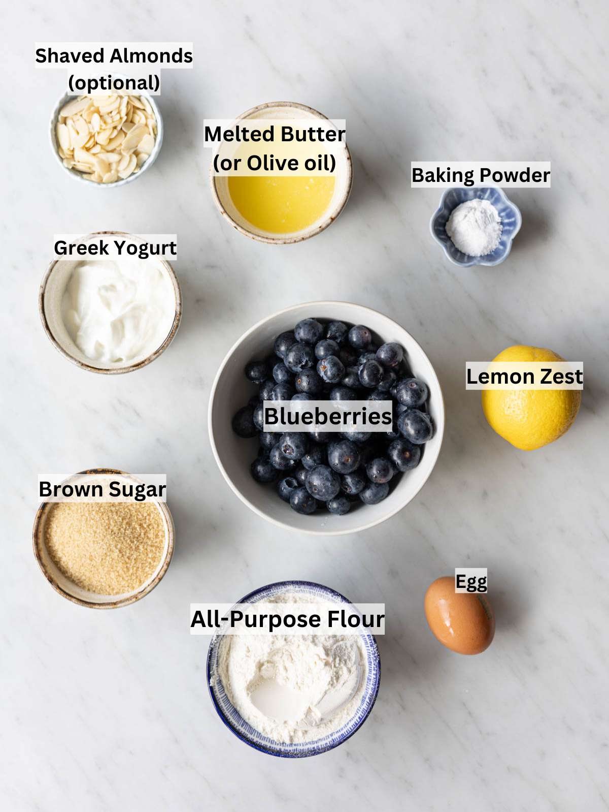 Blueberries, lemon, brown sugar, yogurt, egg, flour, and butter on a marble table.