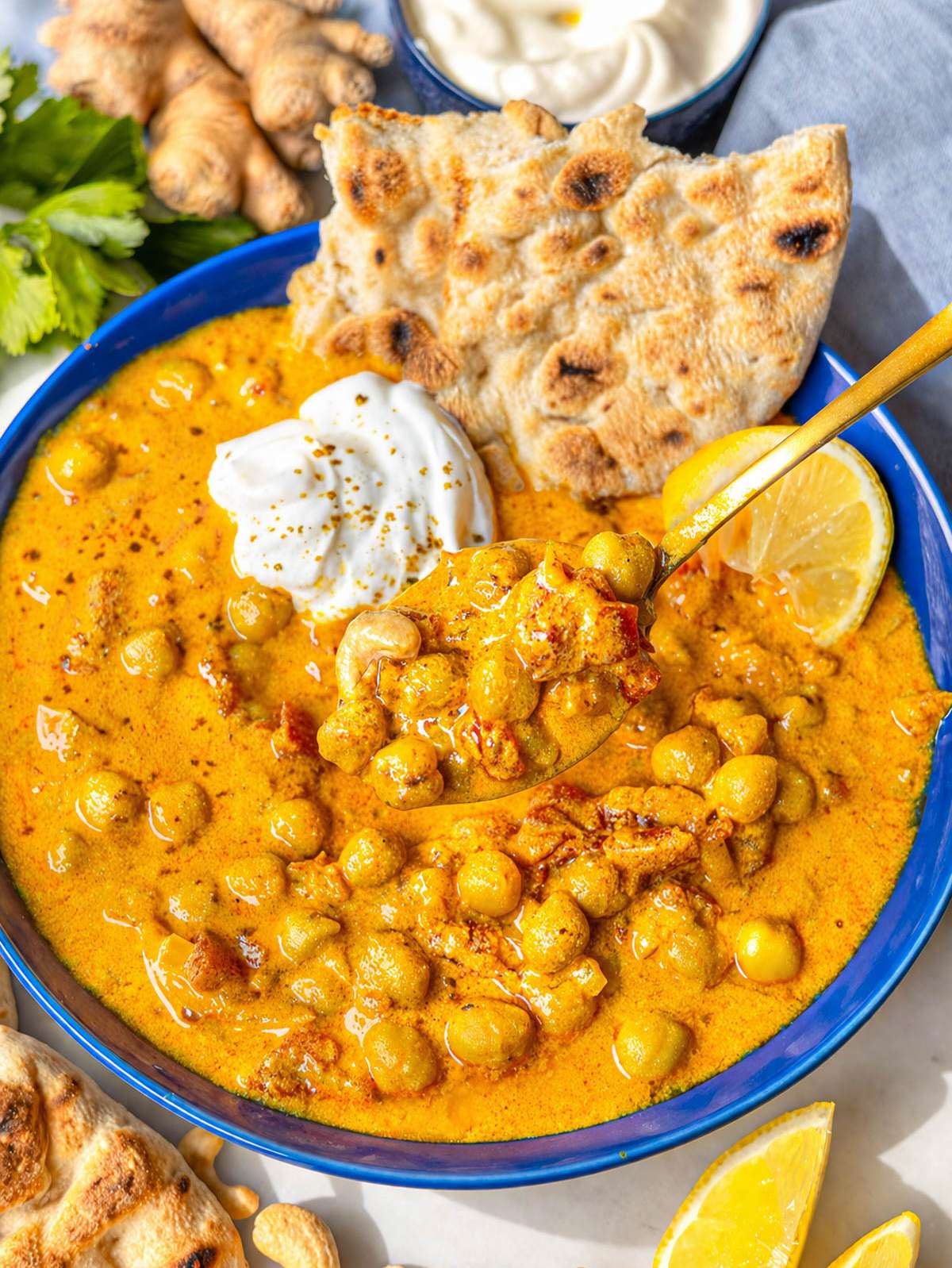 Bowl of Greek yogurt chickpea curry with naan and fresh lemon on marble surface.