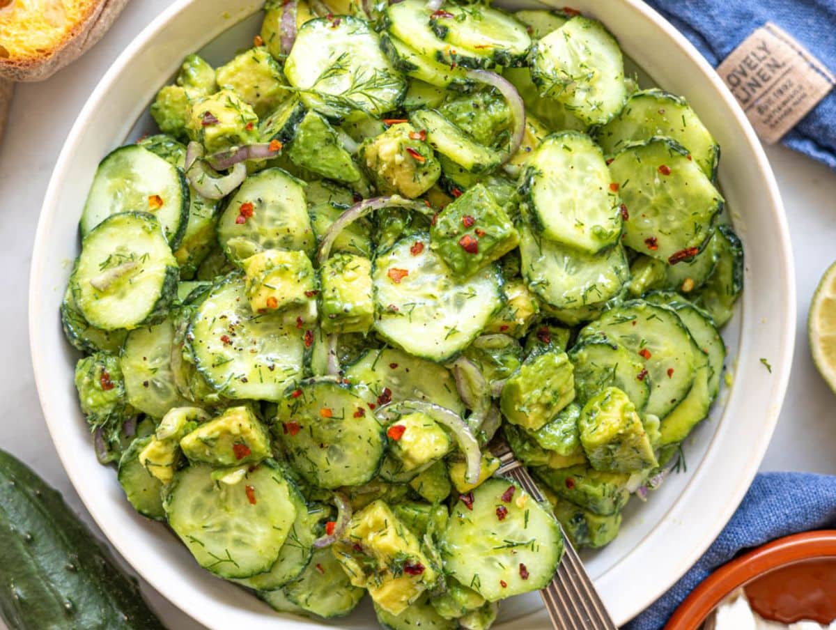 Cucumber avocado salad served in bowl with bread and soft daylight on marble table.