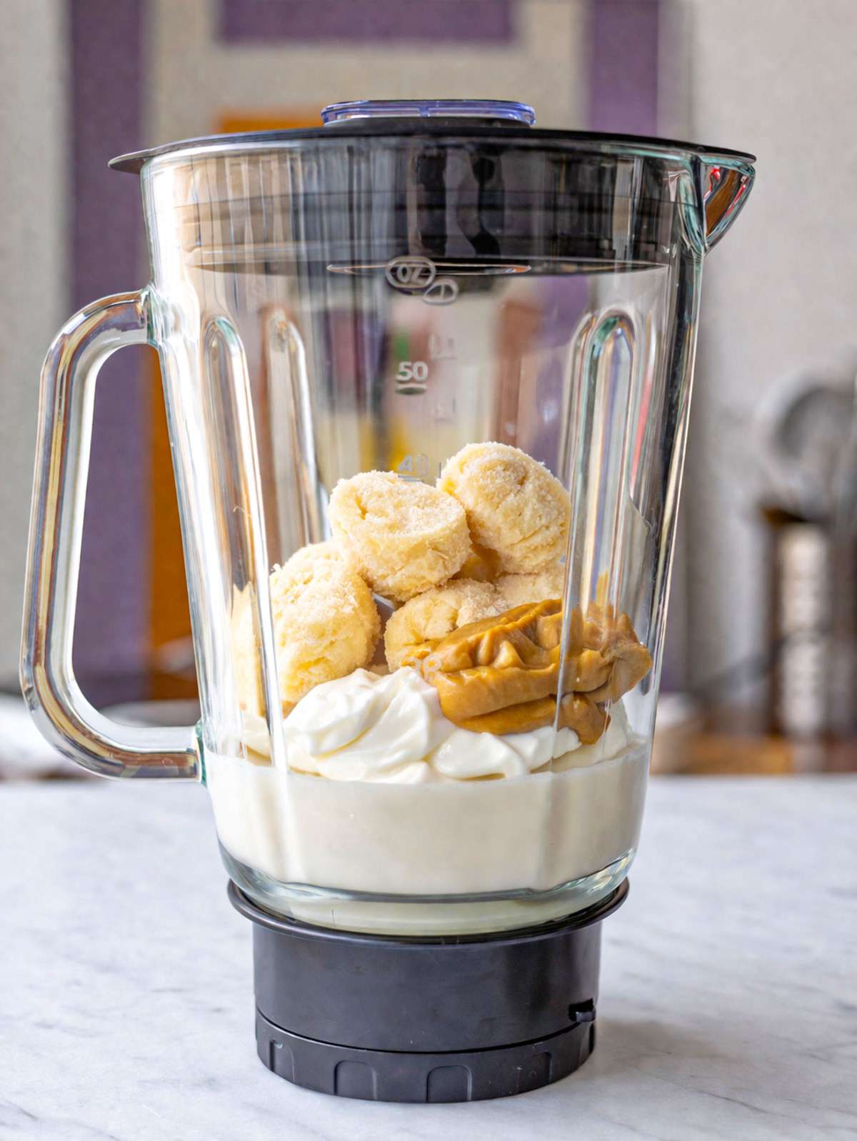 Frozen banana slices, peanut butter, yogurt, and milk layered in blender on marble surface.