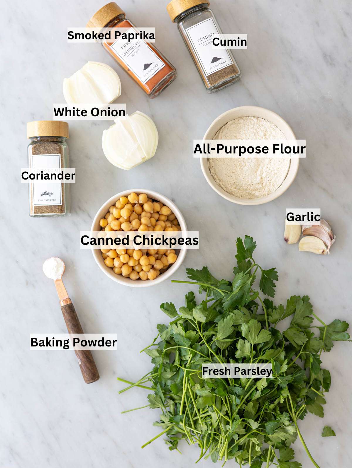 Ingredients arranged on marble surface with chickpeas, herbs, onion, garlic, and spices in natural light.