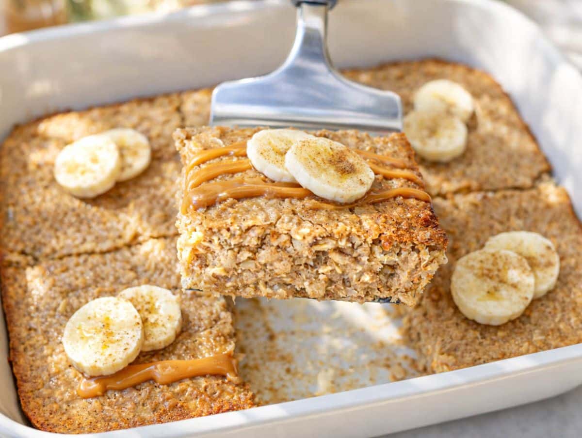 Peanut butter banana baked oatmeal in baking dish with slice lifted and banana and drizzle on top.