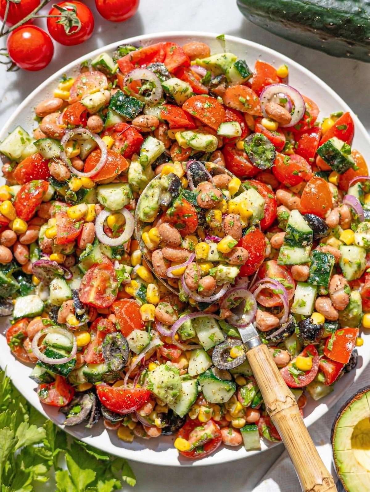 Pinto bean salad in bowl on marble table with avocado, tomatoes, cucumber, and herbs.
