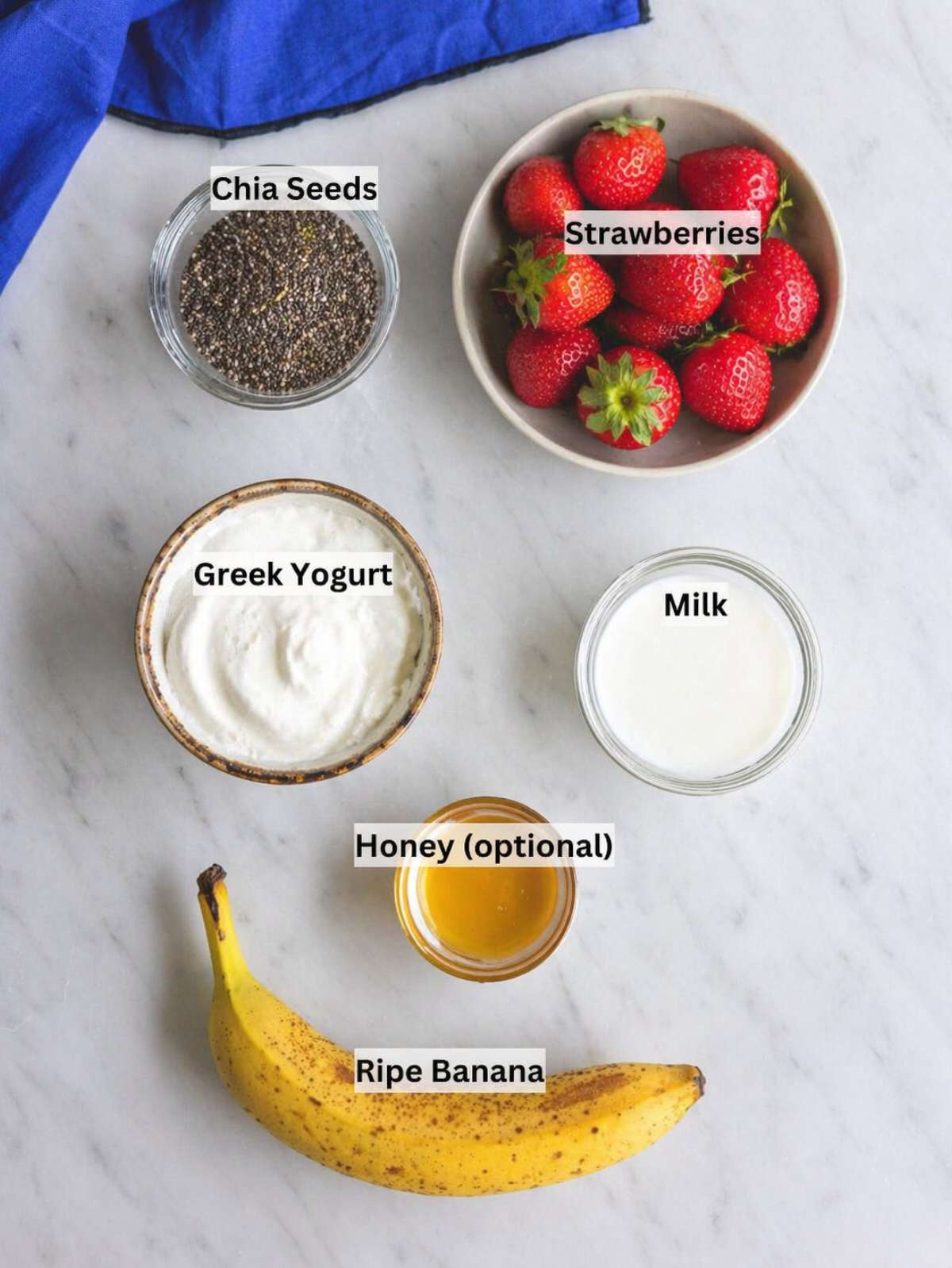 Ripe banana, fresh strawberries, yogurt, milk, chia seeds arranged on marble surface in natural light.