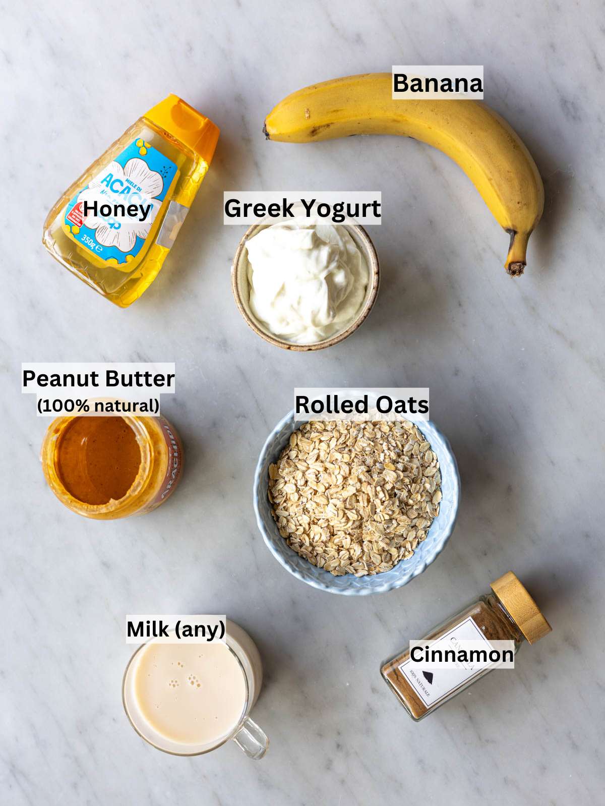 Rolled oats banana peanut butter yogurt milk and honey arranged on marble table in natural light.