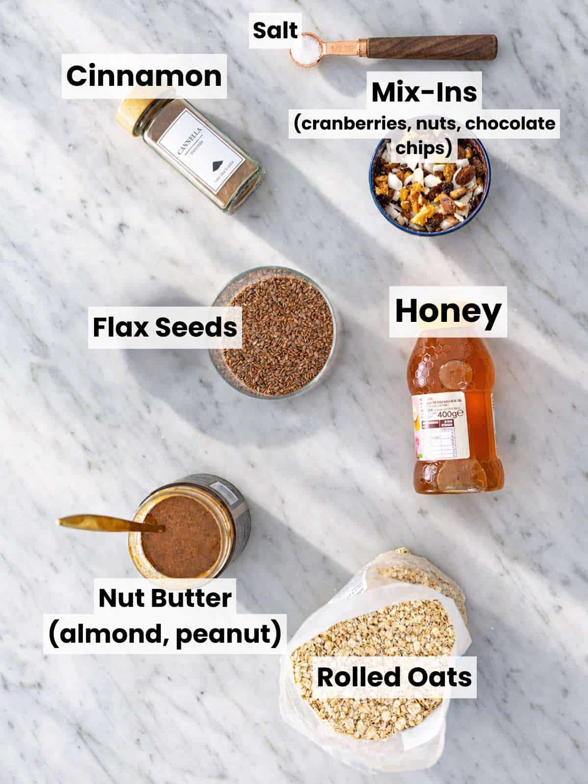 Rolled oats, nut butter, honey, flax seeds, cinnamon, and chocolate chips on marble surface.