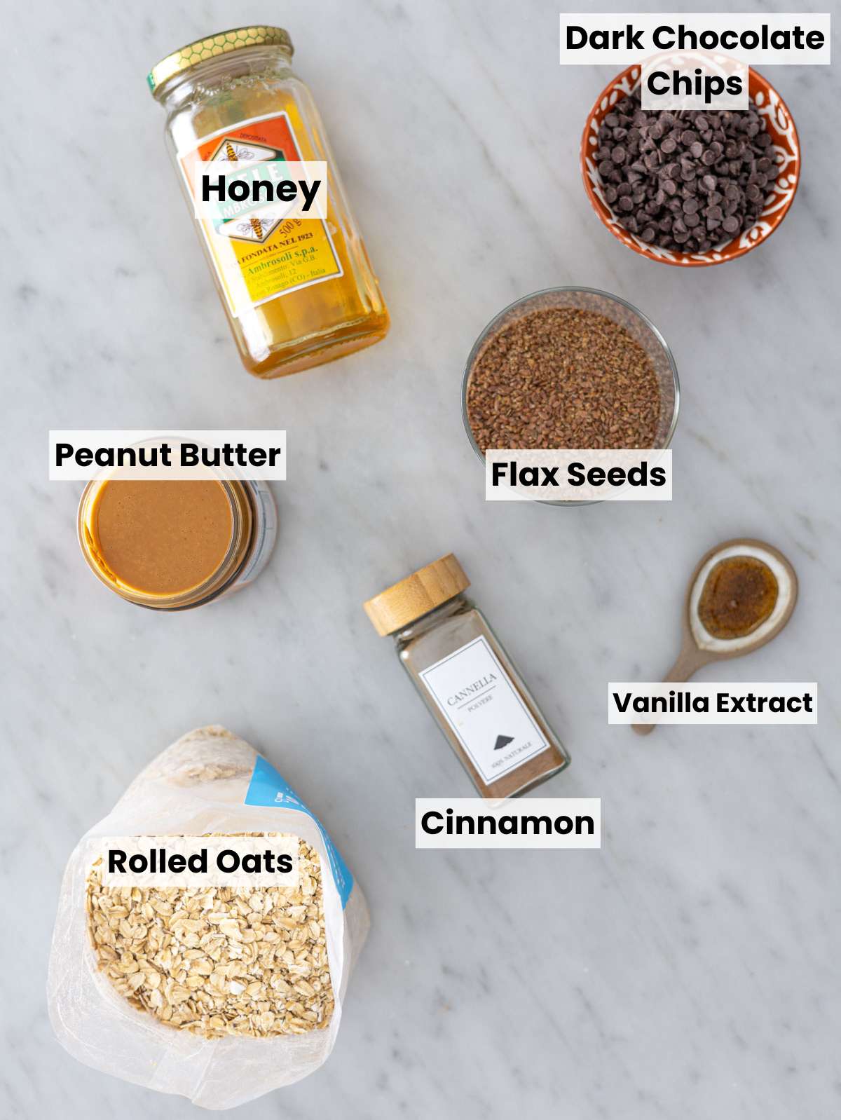 Rolled oats, peanut butter, chocolate chips, honey, and seeds on a marble table.