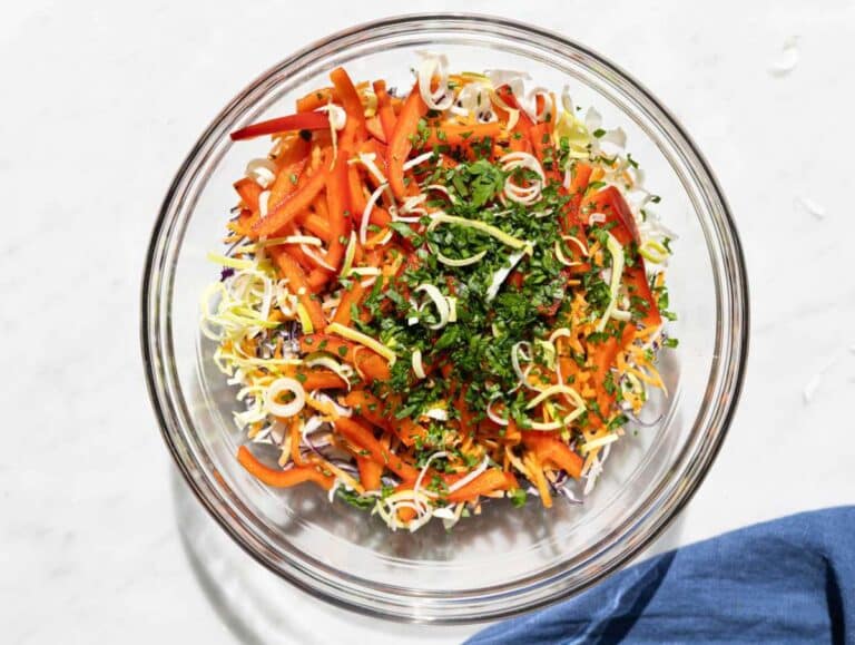 Shredded cabbage, carrots, and herbs combined in a glass bowl under natural light.