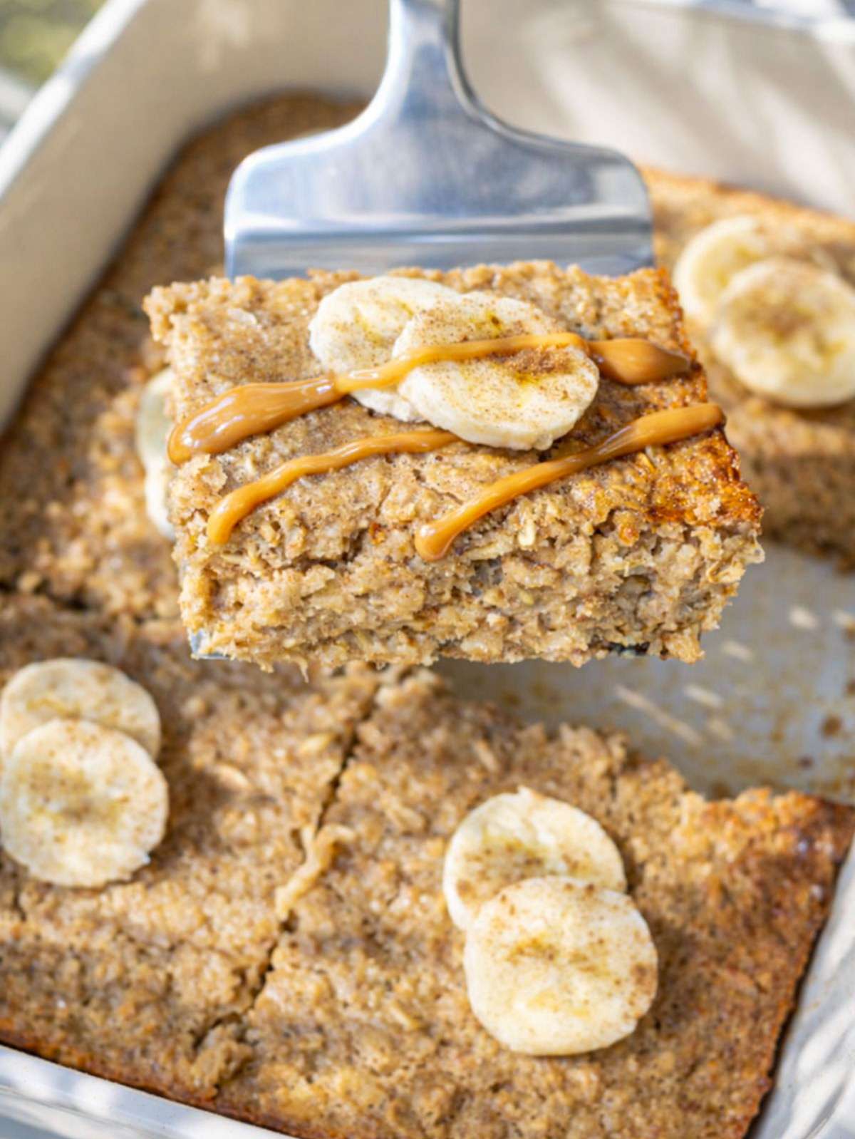 Slice of peanut butter banana baked oatmeal lifted from pan with banana slices and drizzle.