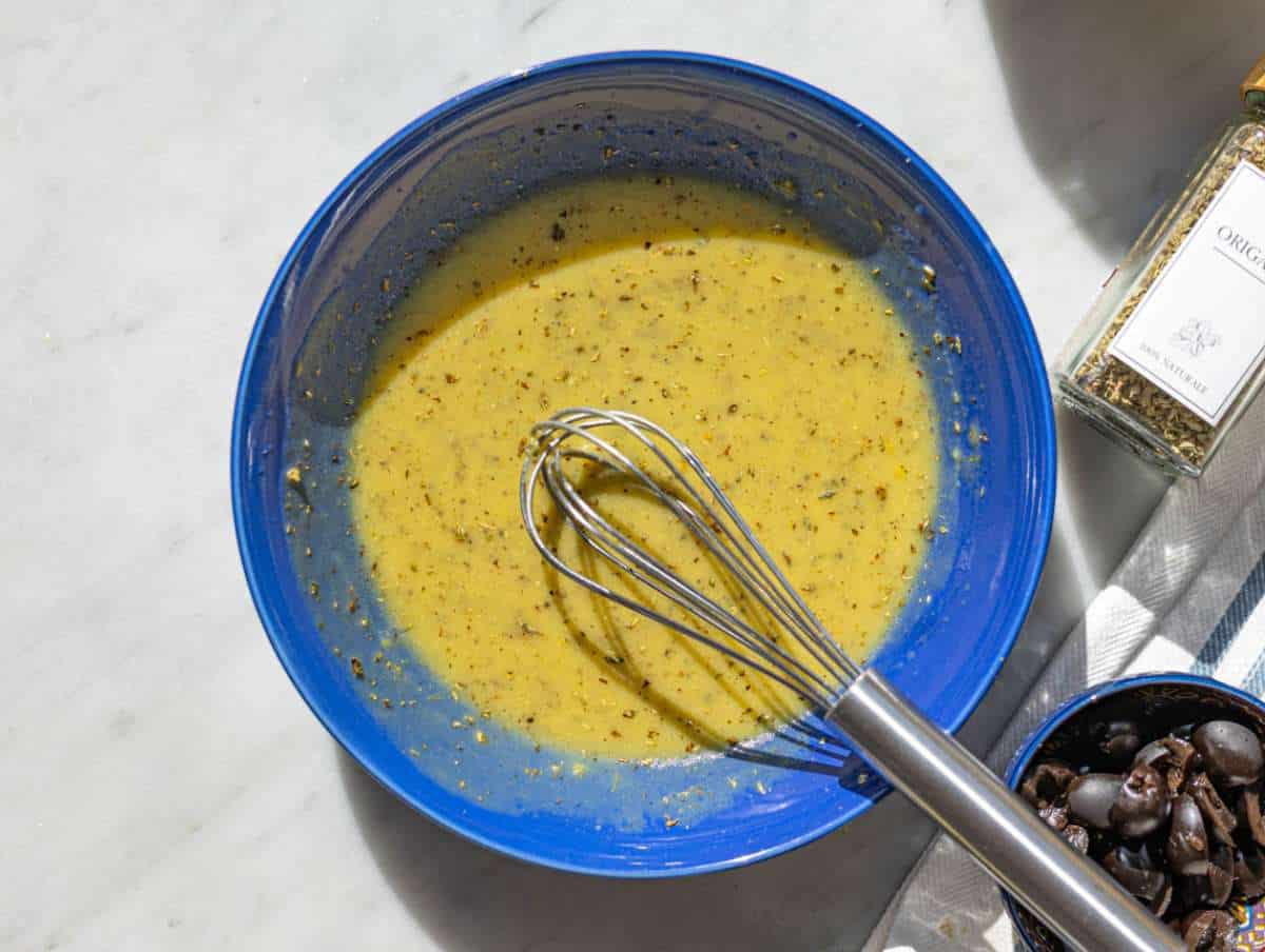 Step 1 Whisking olive oil dressing in bowl with herbs and mustard on marble table.