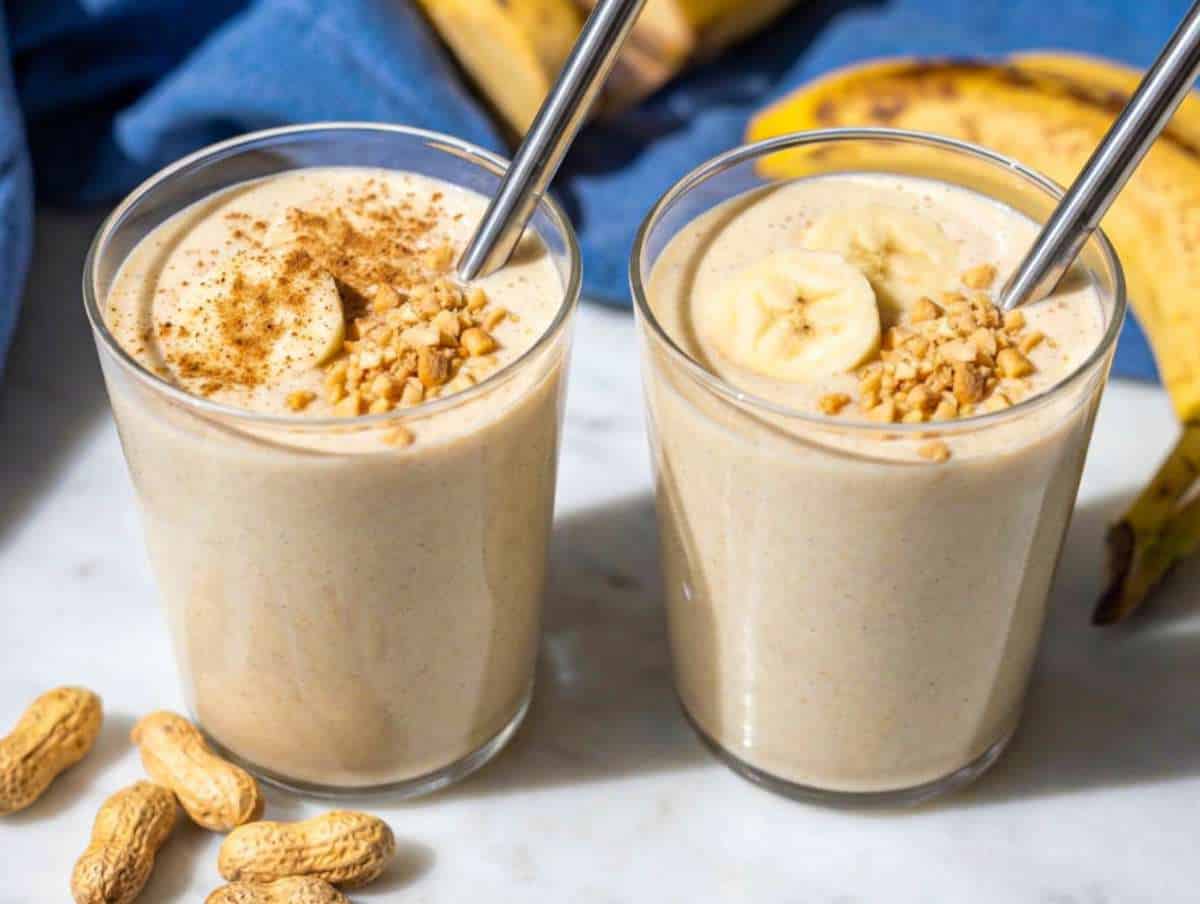 Two creamy banana peanut butter smoothies with metal straws, topped with banana slices and crushed peanuts.