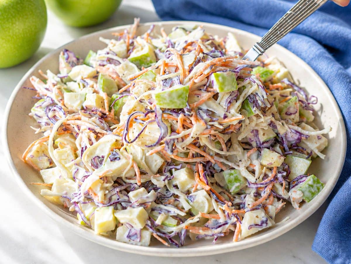 Bowl of apple coleslaw beside whole apples on bright marble surface.