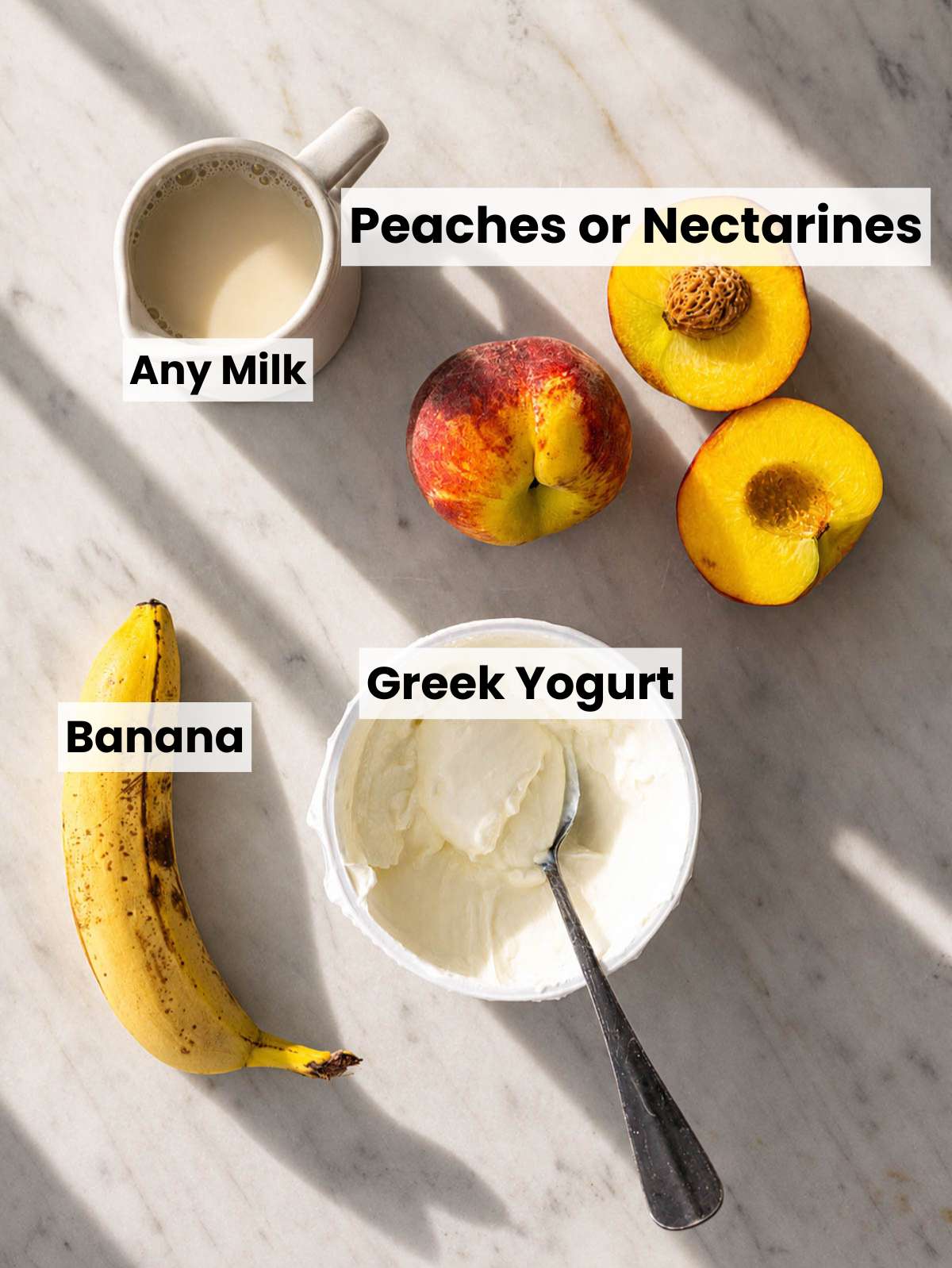 Fresh peaches, Greek yogurt, banana, honey, and ice on a marble counter near window light.
