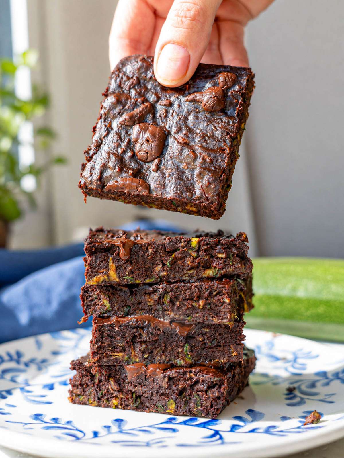 Hand lifting a fudgy zucchini brownie from a stack on a patterned plate.