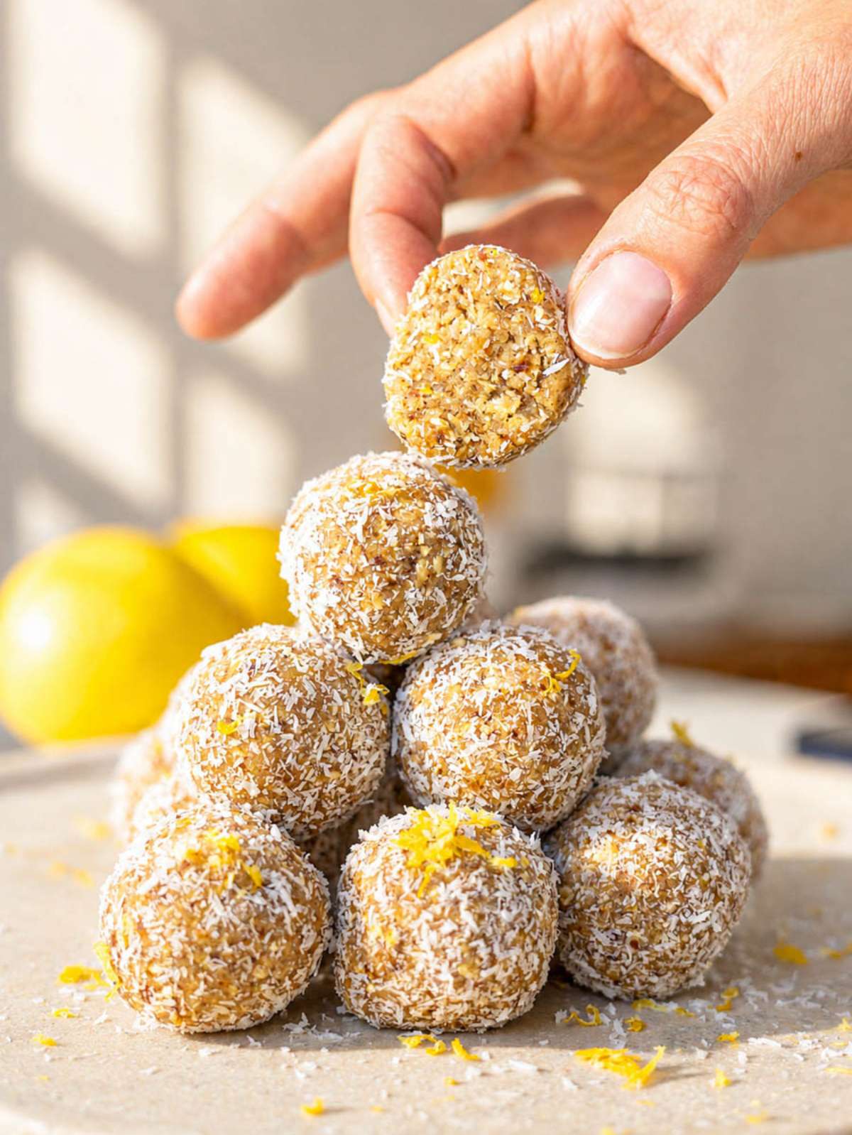 Hand placing coconut-coated Lemon Energy Ball onto stacked bites beside fresh lemons in natural window light.
