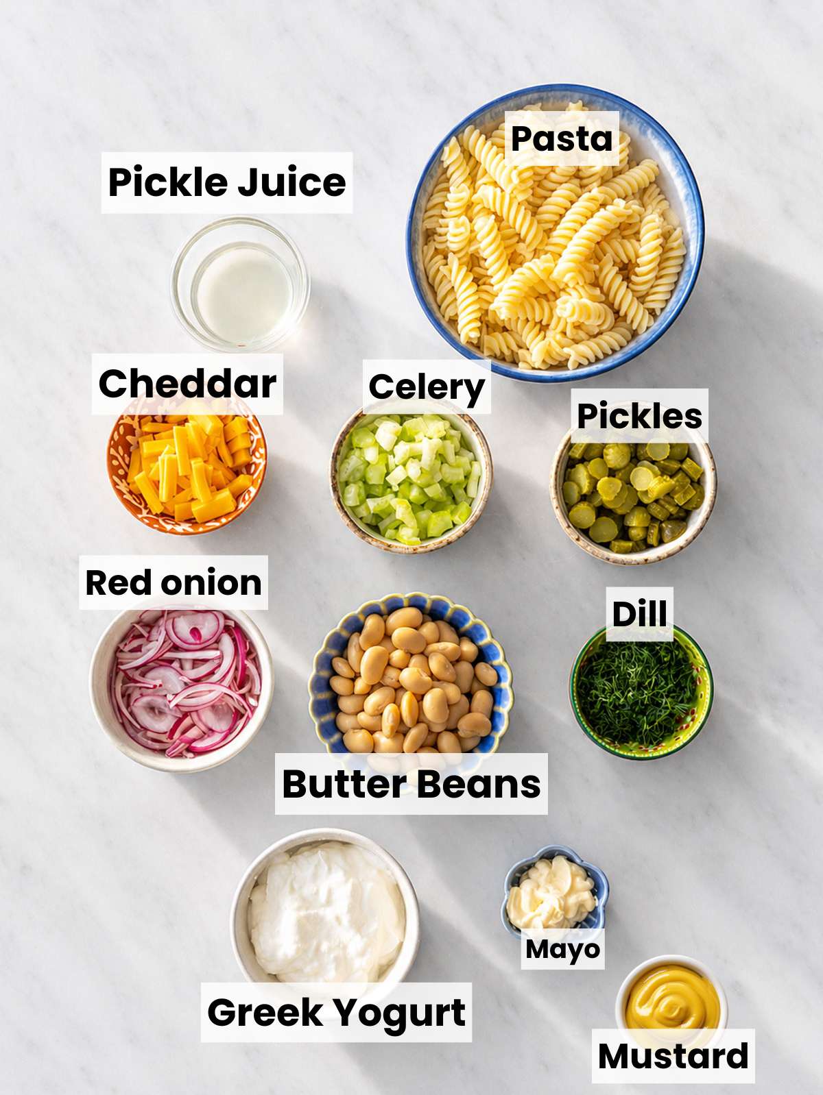 Ingredients for healthy dill pickle pasta salad arranged in bowls on white marble countertop.