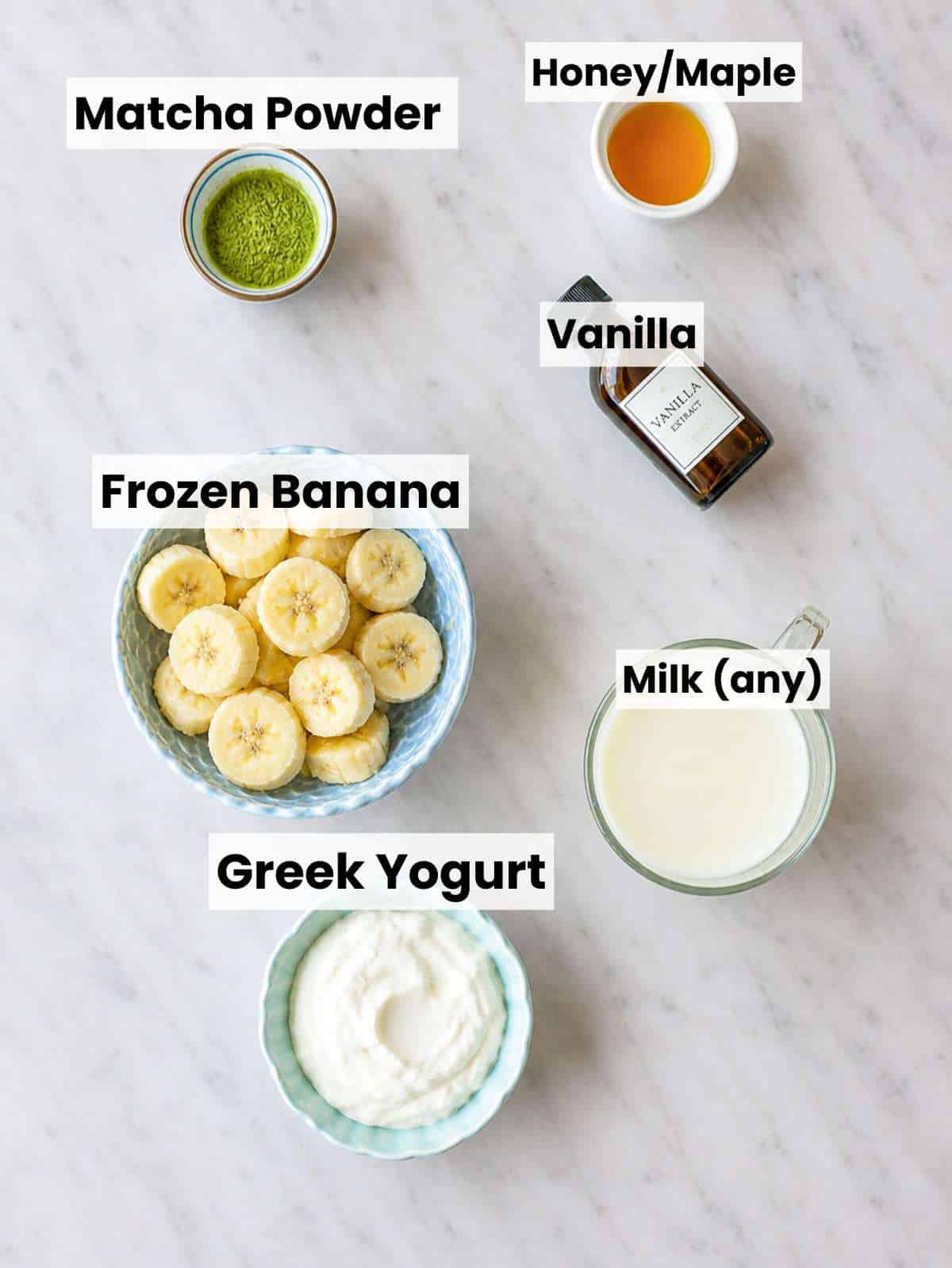 Matcha powder, banana, Greek yogurt, almond milk, honey arranged on marble in natural light.