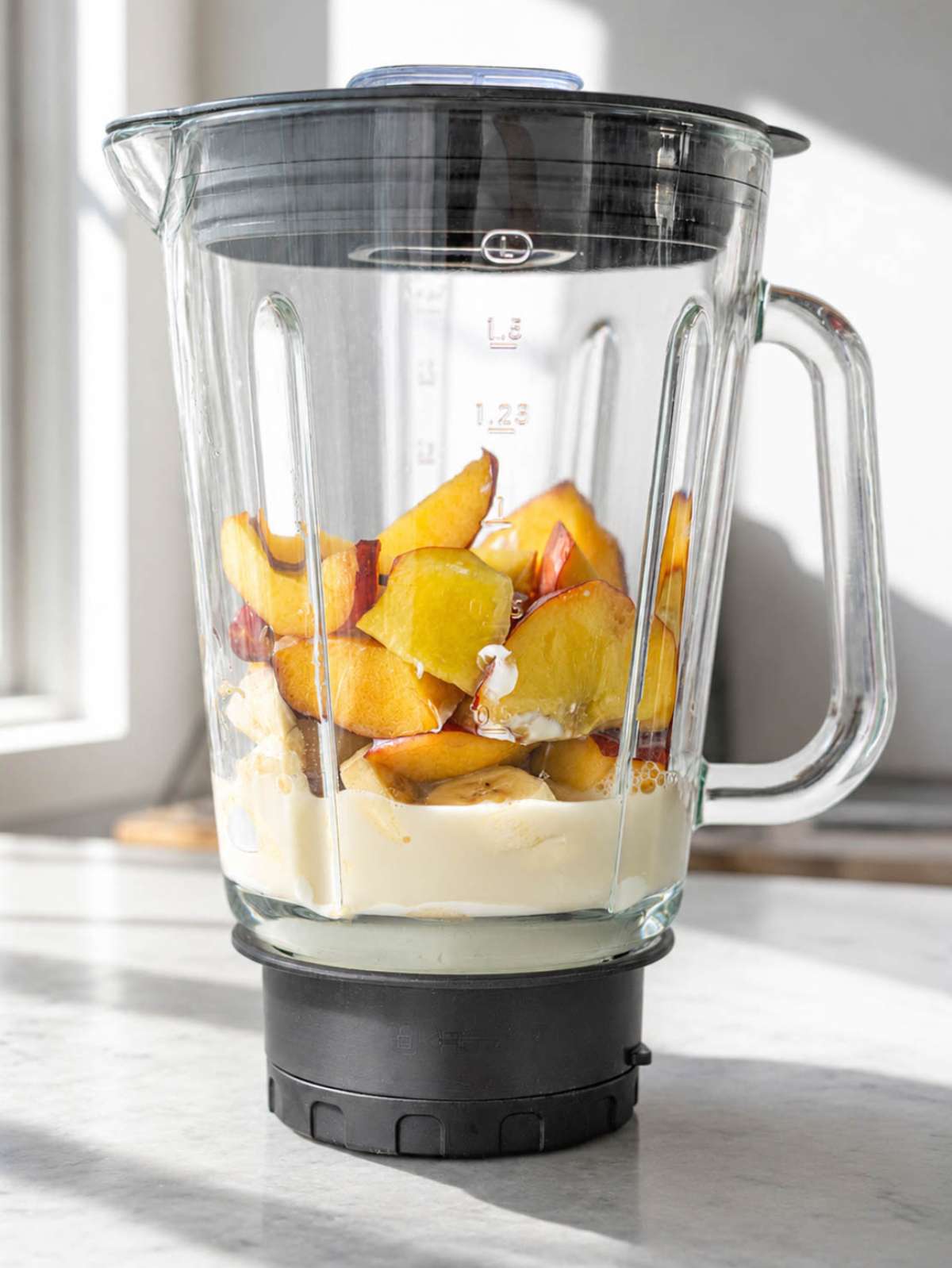 Peach slices, banana, and Greek yogurt layered in a blender on a marble countertop near sunlight.