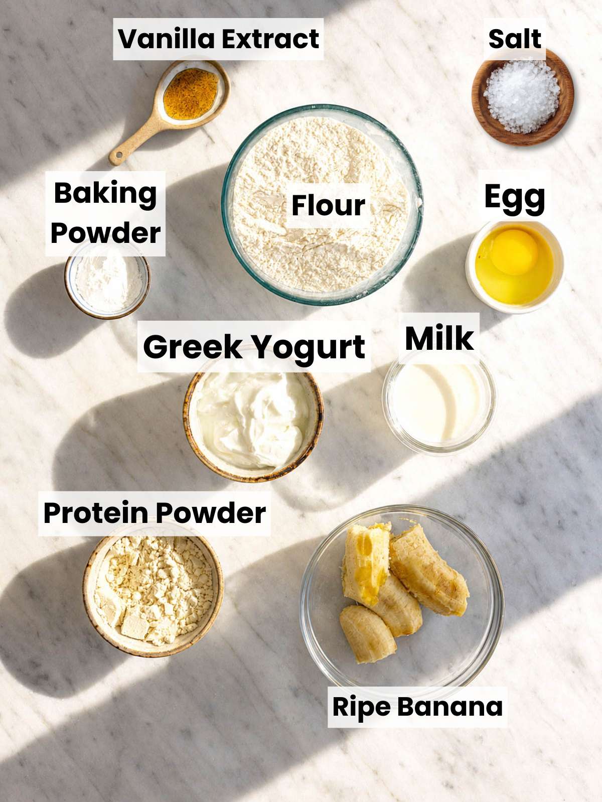 Ripe bananas, Greek yogurt, eggs, flour, and protein powder on white marble counter.