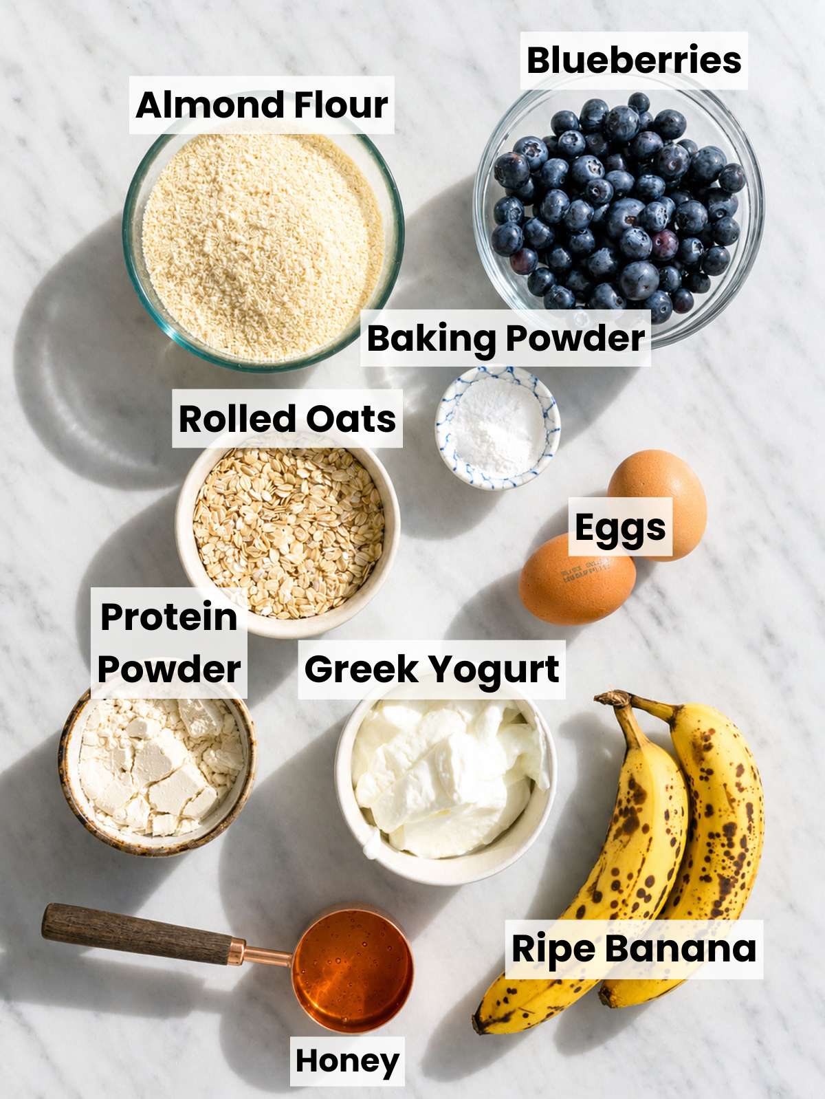 Ripe bananas, blueberries, oats, Greek yogurt, and protein powder on a marble counter.