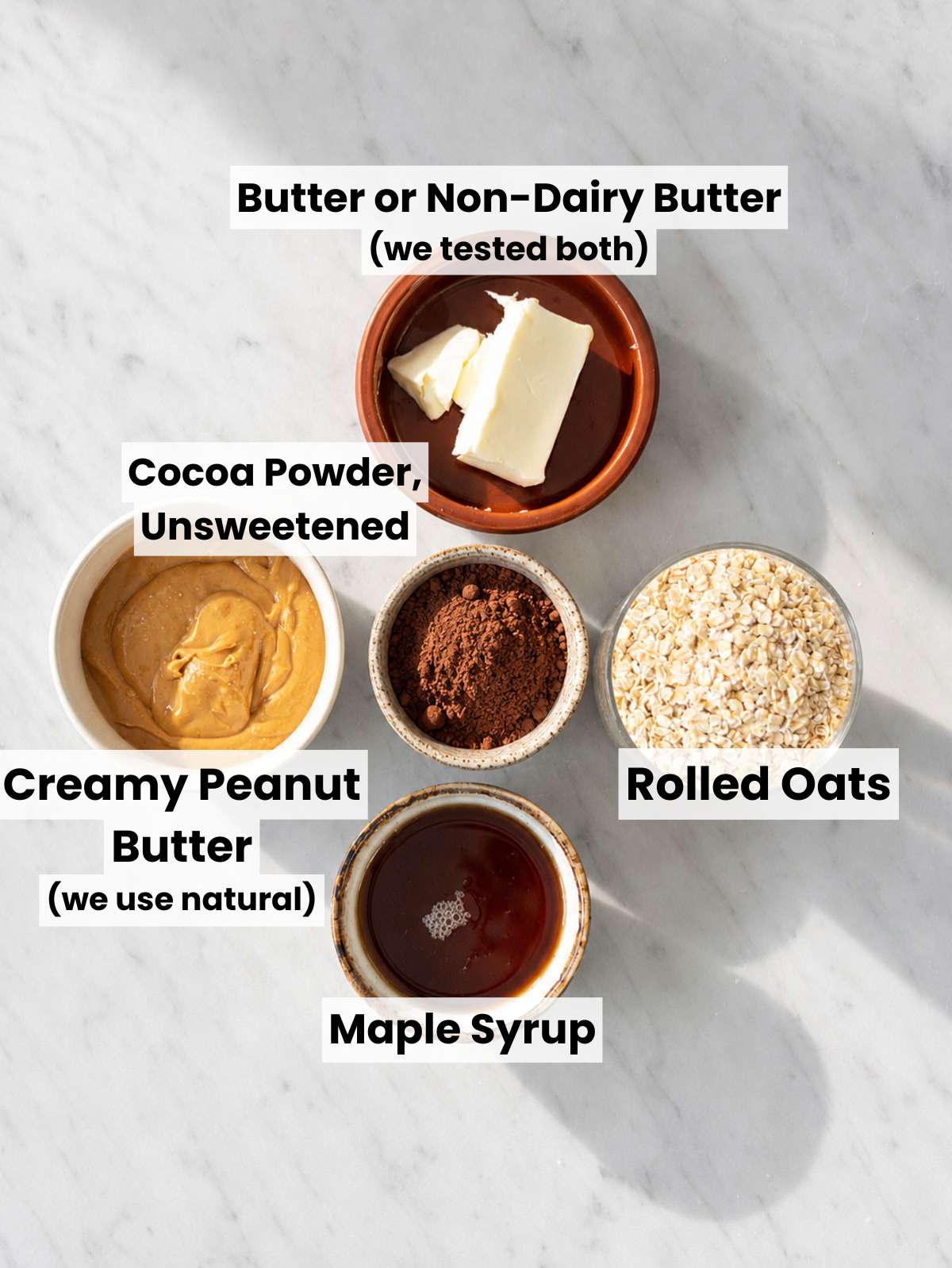 Rolled oats, cocoa, peanut butter, maple syrup, butter, vanilla, and salt on marble.