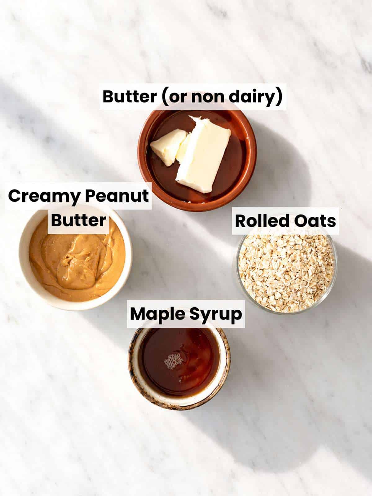 Rolled oats, peanut butter, maple syrup, butter, and vanilla on marble surface.