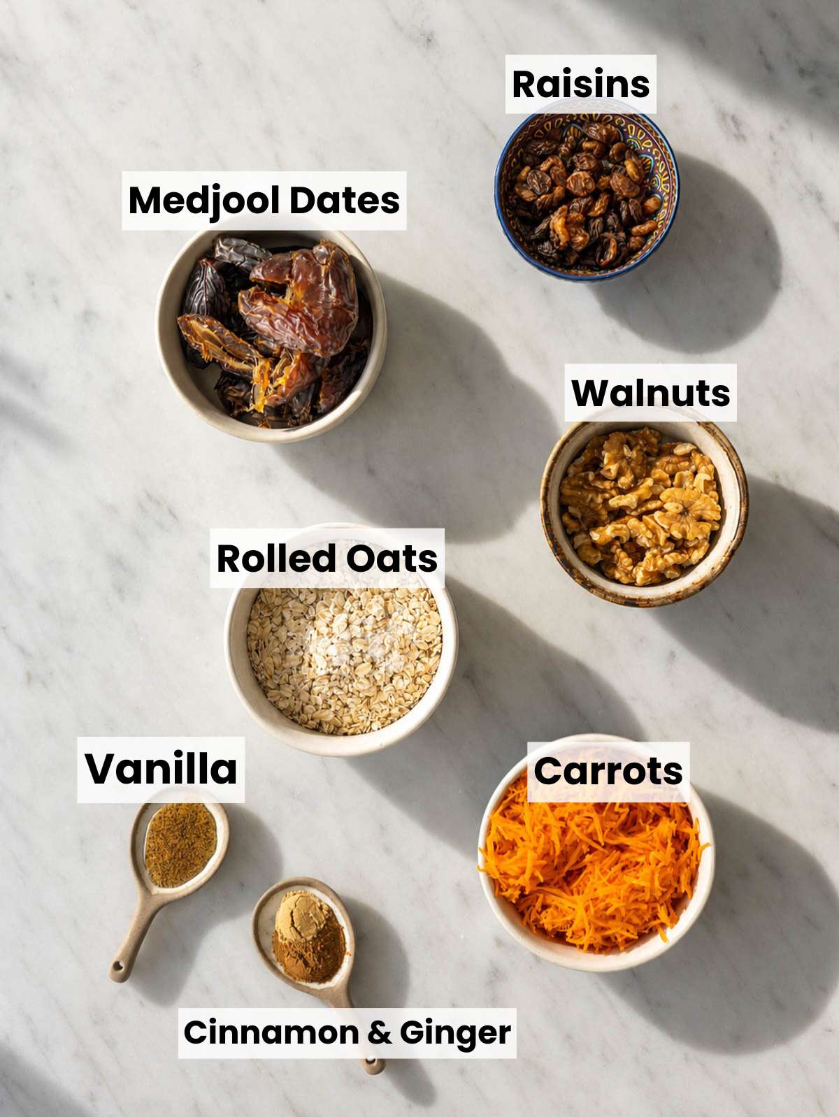 Rolled oats, shredded carrots, walnuts, dates, and spices on white marble counter.
