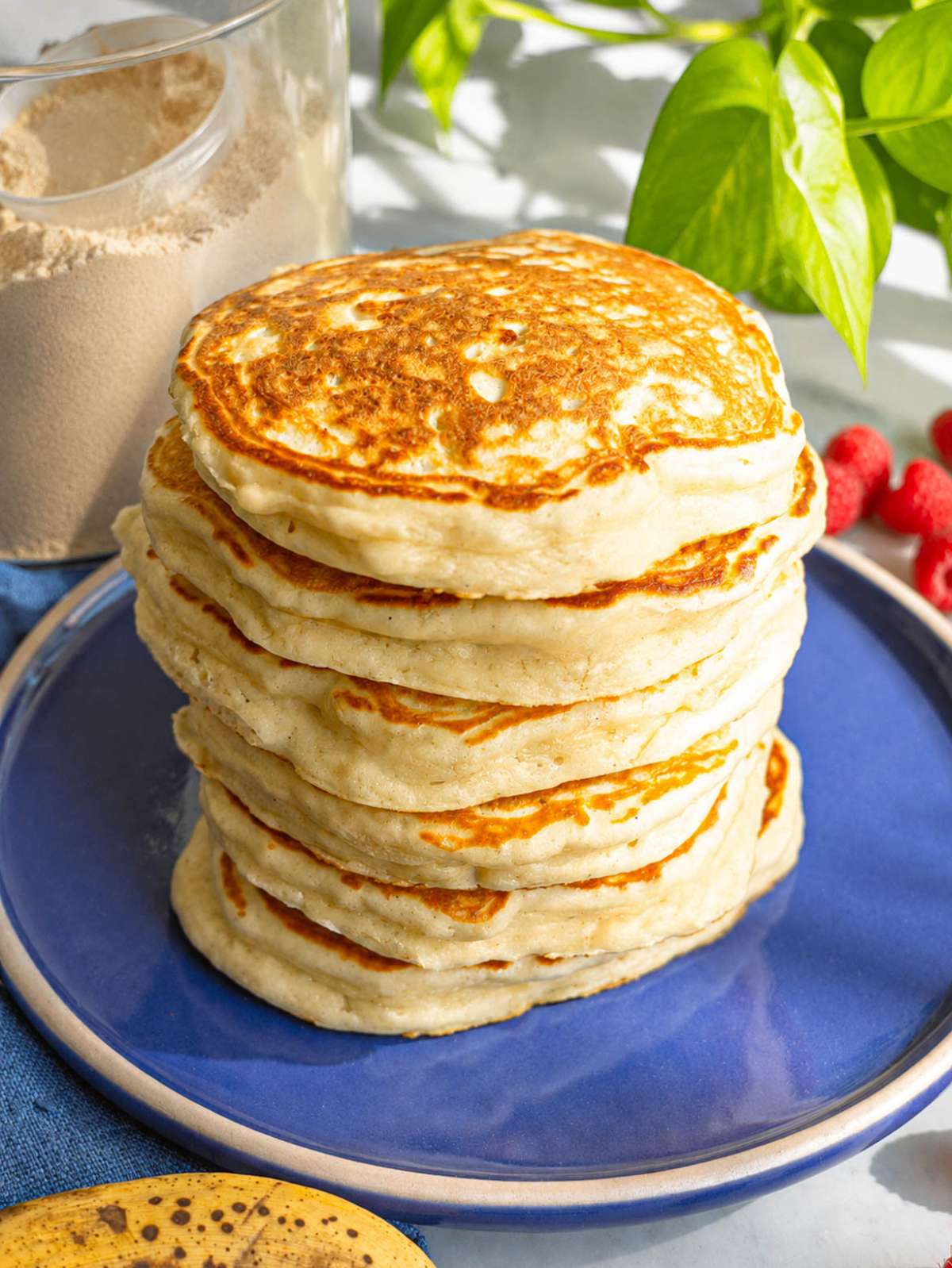 Stack of fluffy protein banana pancakes on blue plate with raspberries and protein powder nearby.