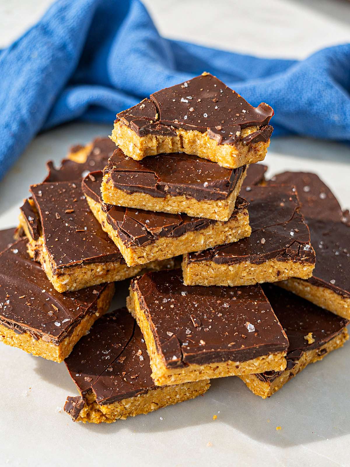 Stacked chocolate peanut butter bars with flaky salt on marble countertop near bright window light.