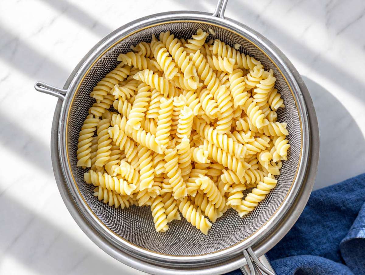 Step 1 Boiled pasta draining in colander with steam rising in natural window light.