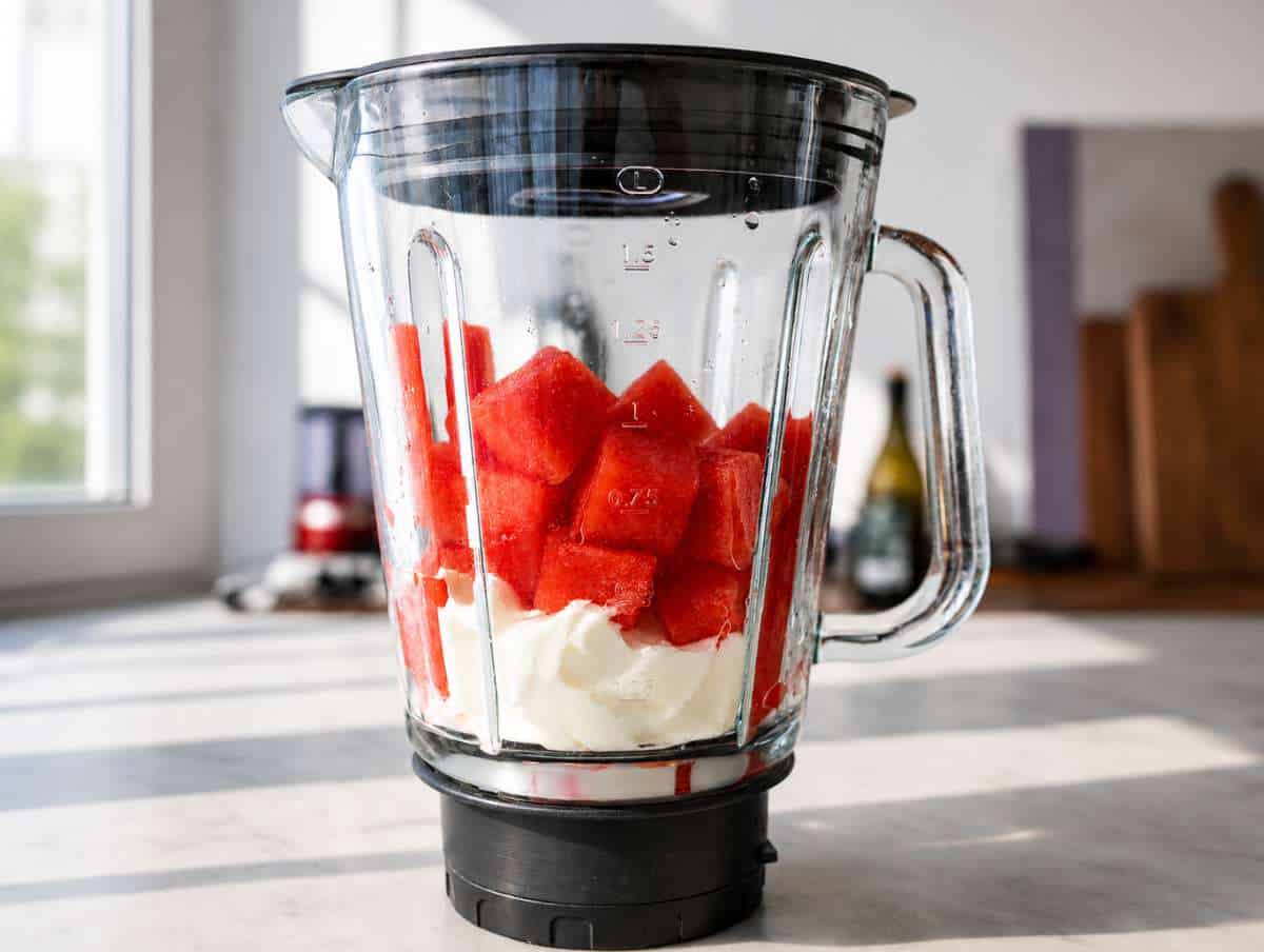 Blender with watermelon smoothie ingredients next to a bright window.