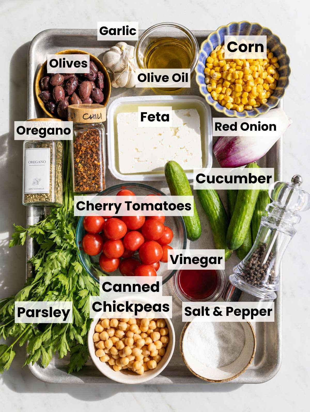 Chickpeas, parsley, cucumber, tomatoes, feta, corn, red onion, and chimichurri ingredients on marble surface.