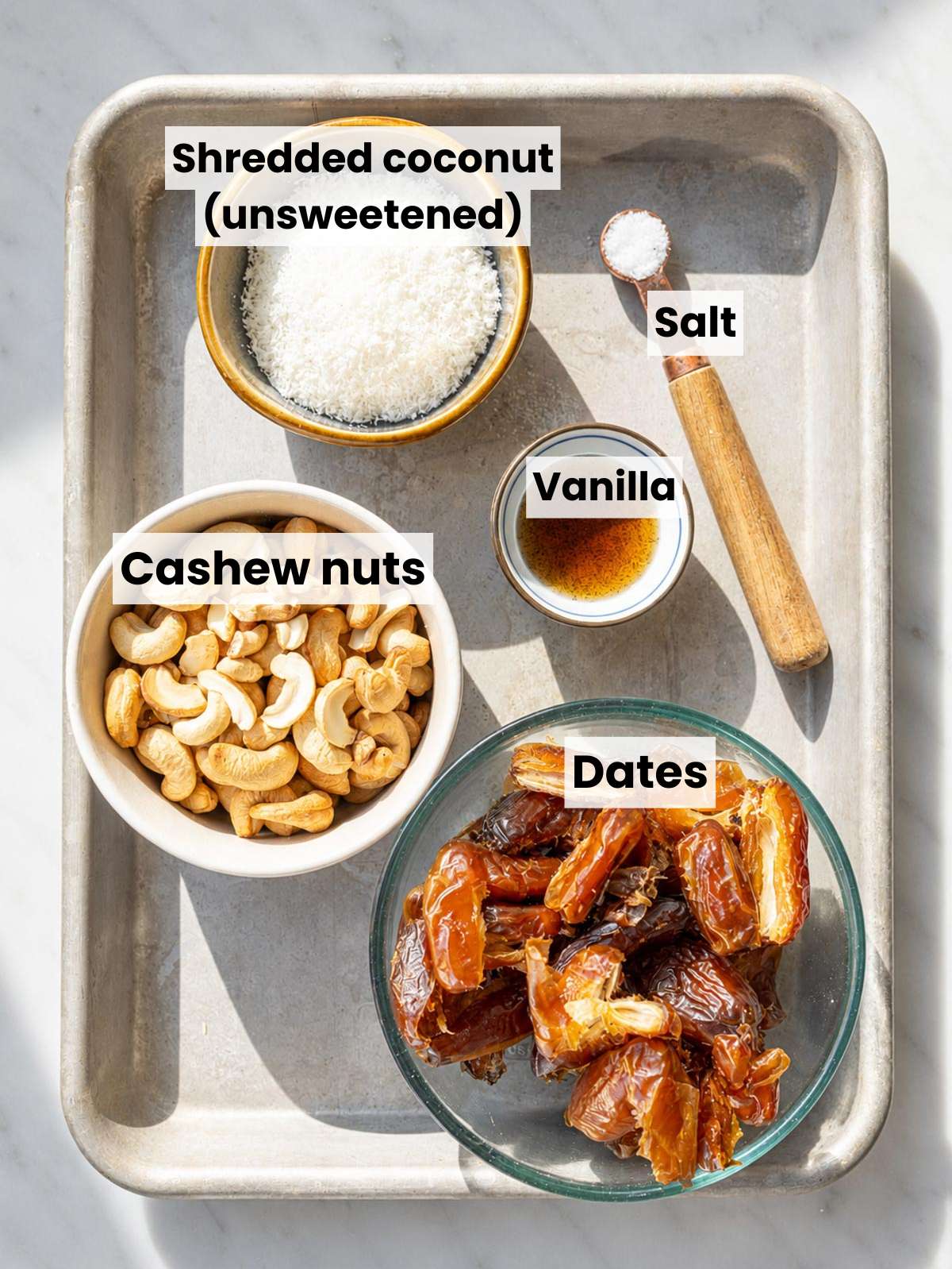 Cashews, dates, coconut, vanilla, and salt on white marble in daylight.