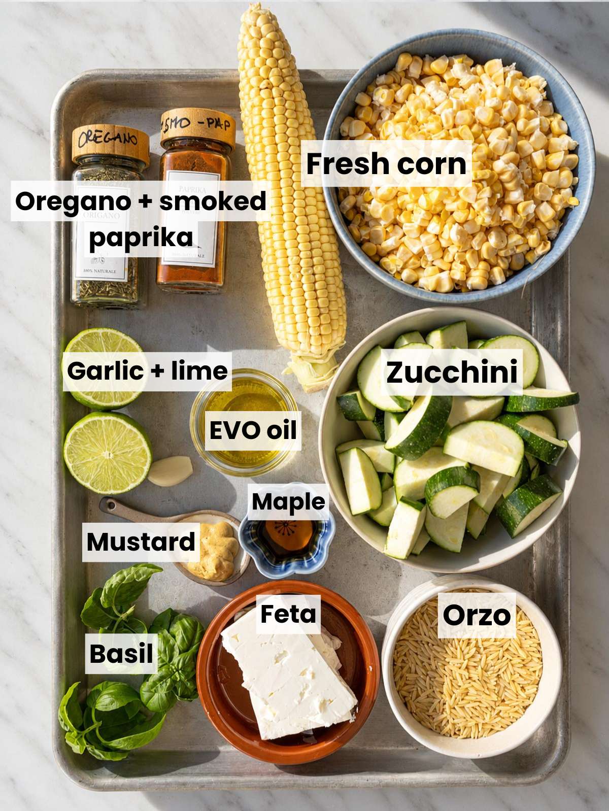 Fresh corn, zucchini, orzo, feta, lime, herbs, and seasonings on white marble.
