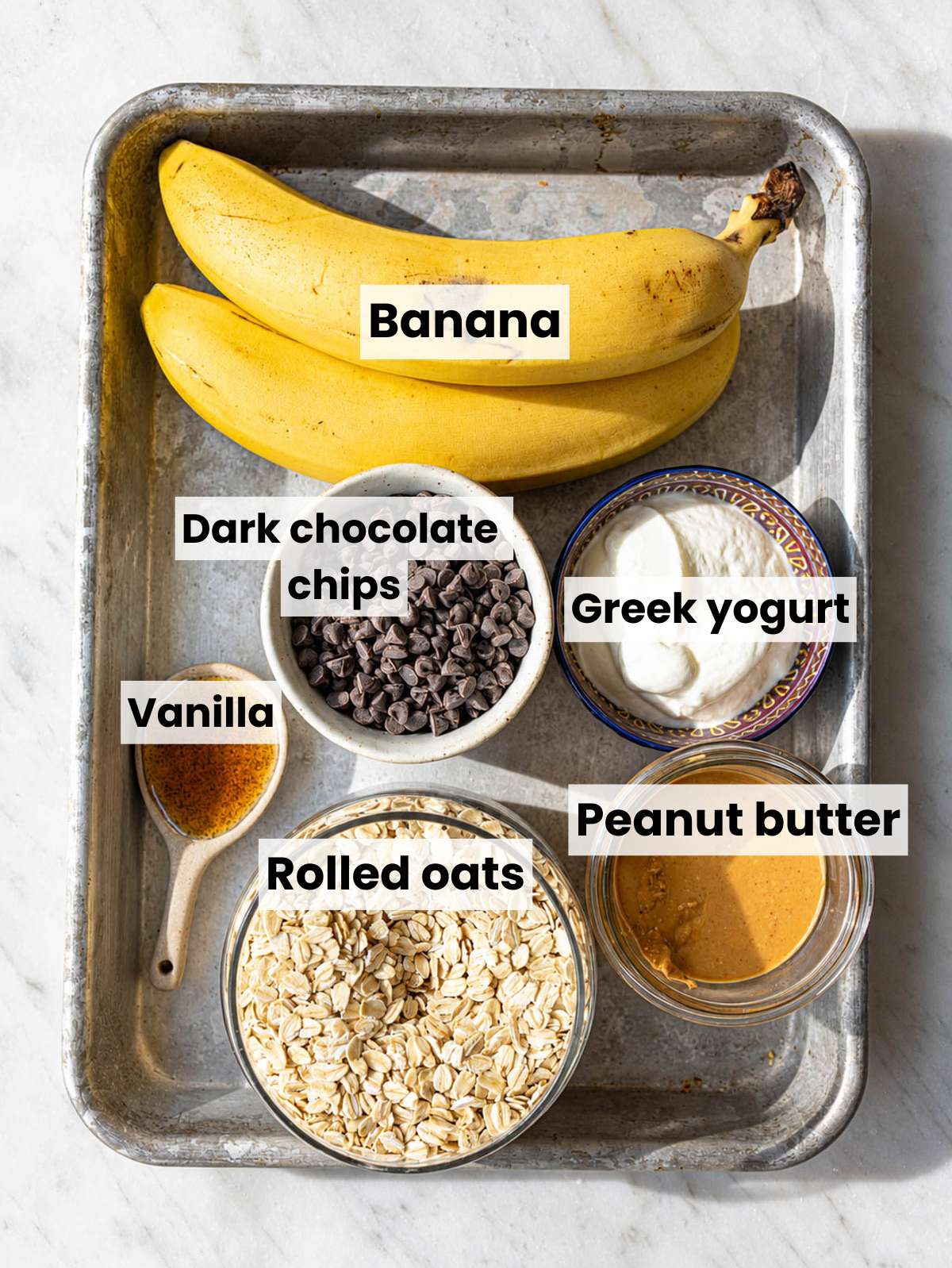 Ripe bananas, Greek yogurt, oats, peanut butter, vanilla, chocolate chips on marble countertop.