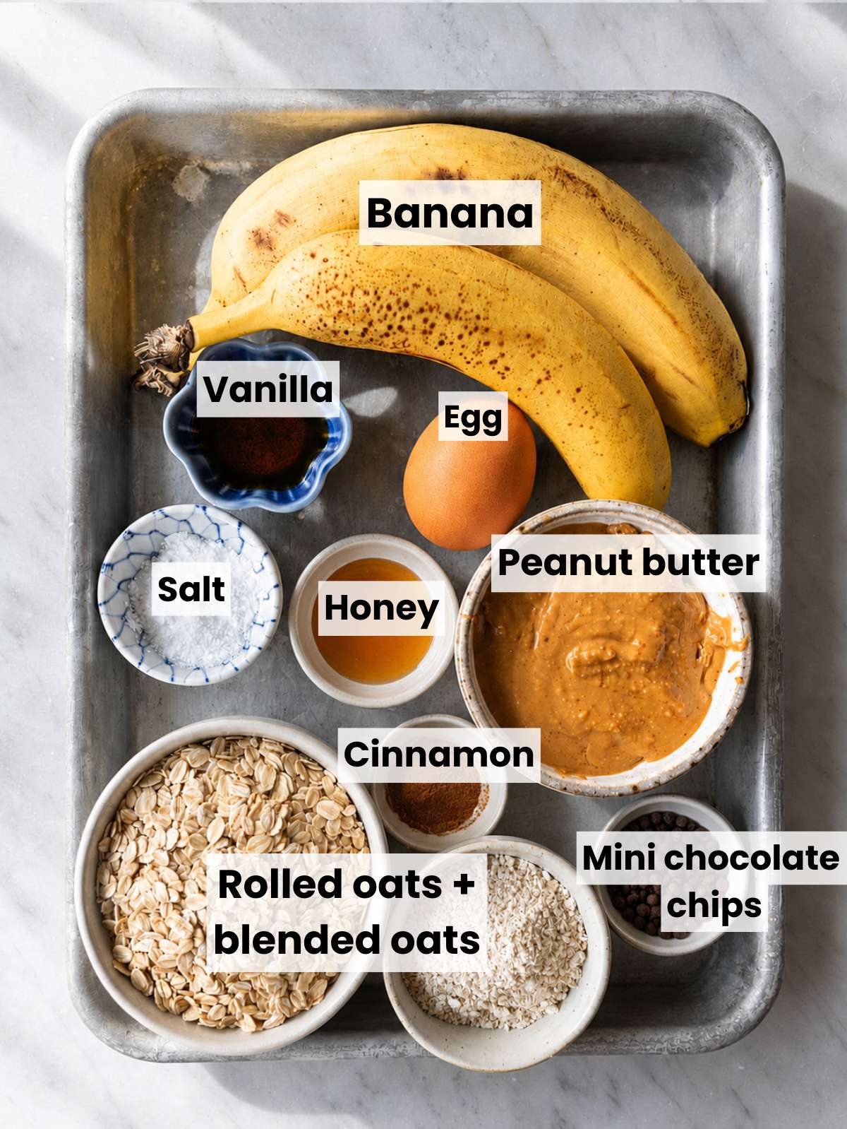 Ripe bananas, peanut butter, oats, honey, egg, chocolate chips on white marble.