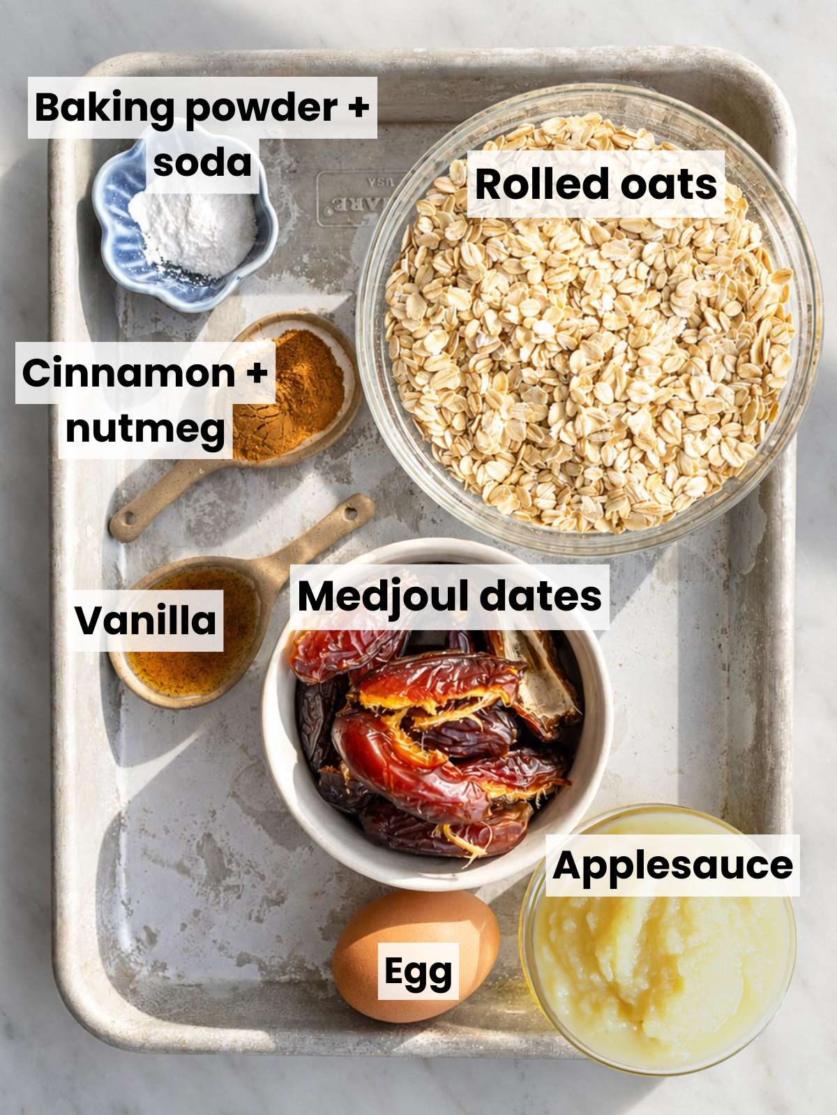 Rolled oats, oat flour, dates, egg, applesauce, spices on white marble countertop.