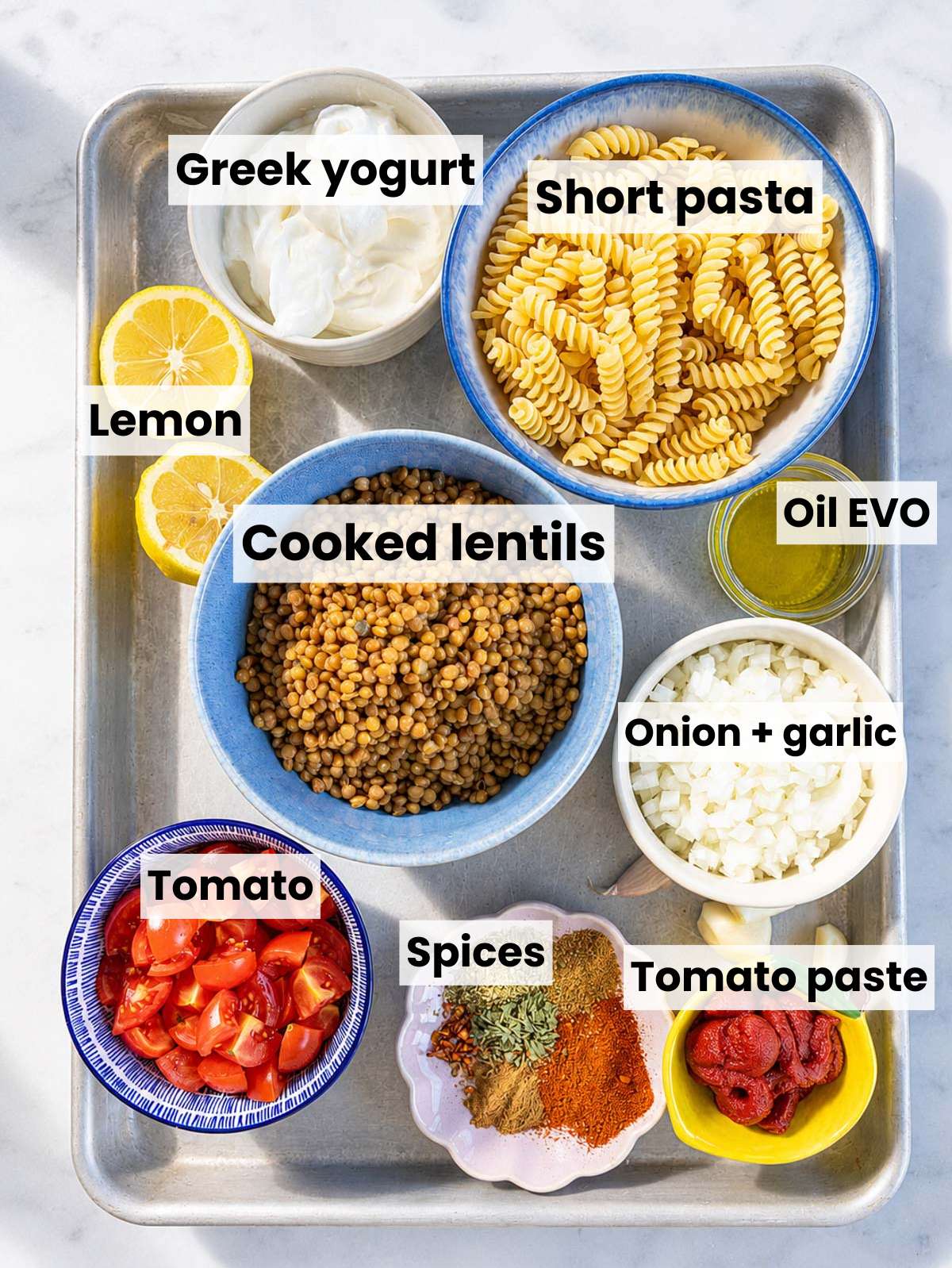 Short pasta, lentils, vegetables, spices, yogurt, and lemon on white Carrara marble.