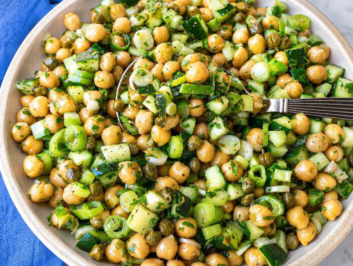 Spoon lifting glossy chickpea salad with cucumber and capers in daylight.